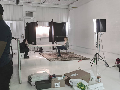 a picture of performers in white studio shooting with lights sound blankets table chairs carpet apple boxes